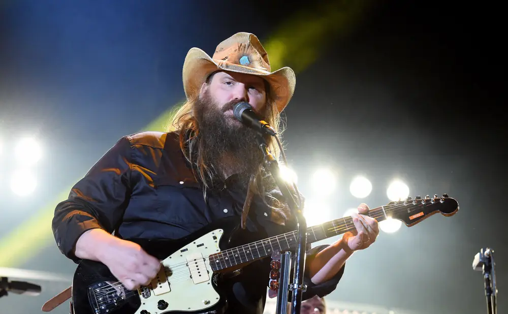 A male singer in a cowboy hat plays a guitar on stage with bright lights behind him.
