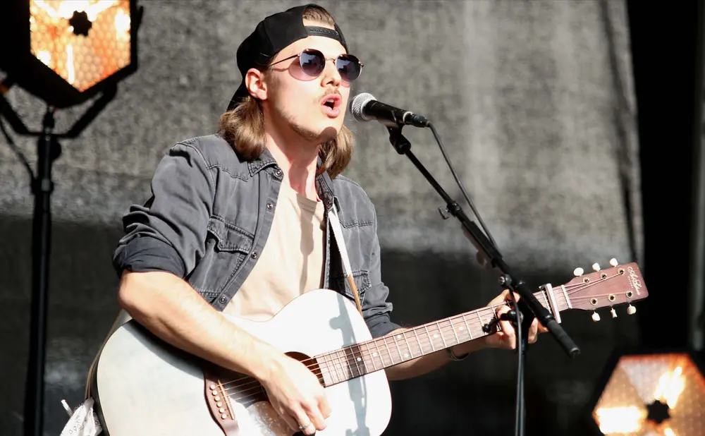 A male musician with sunglasses and a backward baseball cap plays an acoustic guitar and sings into a microphone on stage.