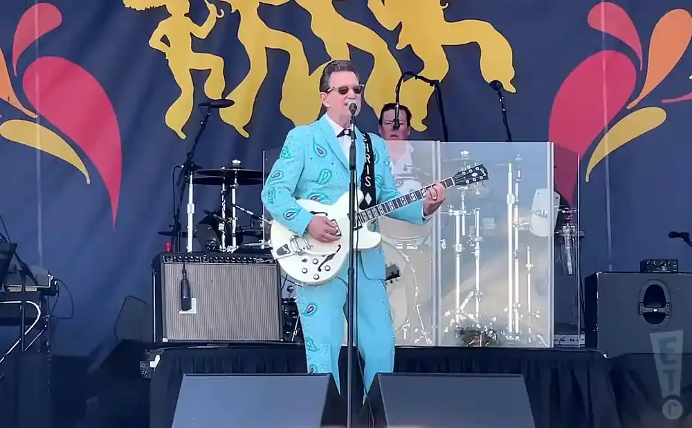 A man in a sparkly blue suit plays a white guitar on stage with a colorful backdrop.