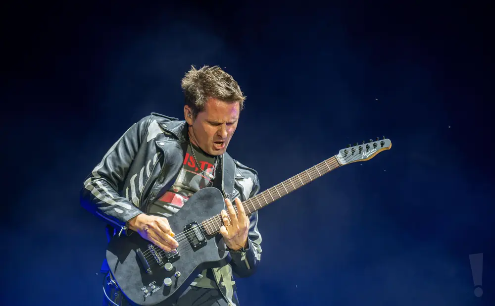 A male guitarist plays a black electric guitar on a dark blue stage.