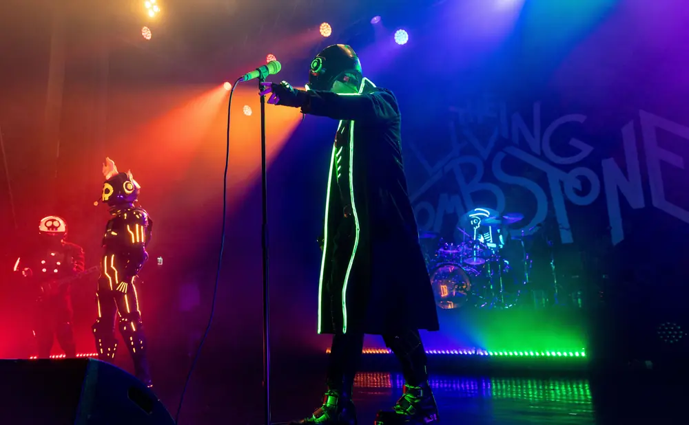 Musicians in futuristic neon costumes perform on stage with dramatic lighting and a band logo.