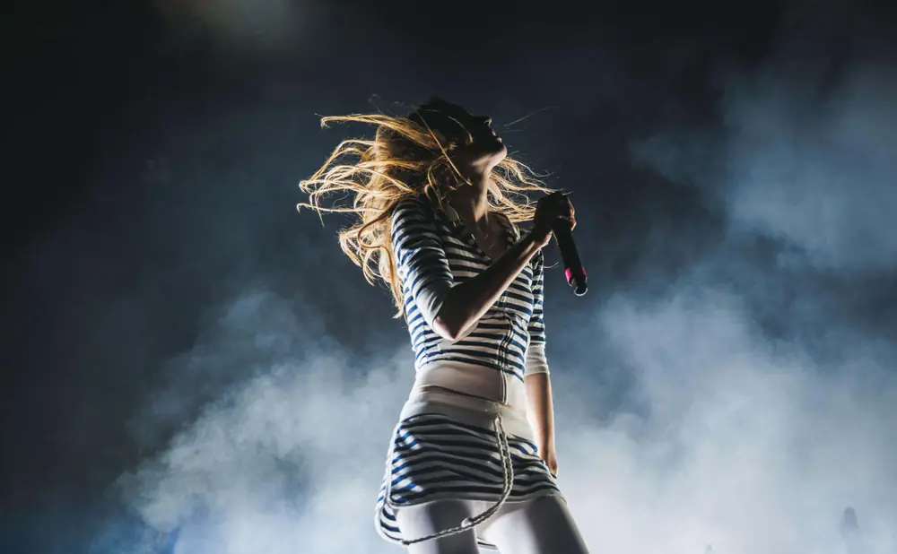 Singer with blonde hair in a striped outfit performs on stage with smoke effects.