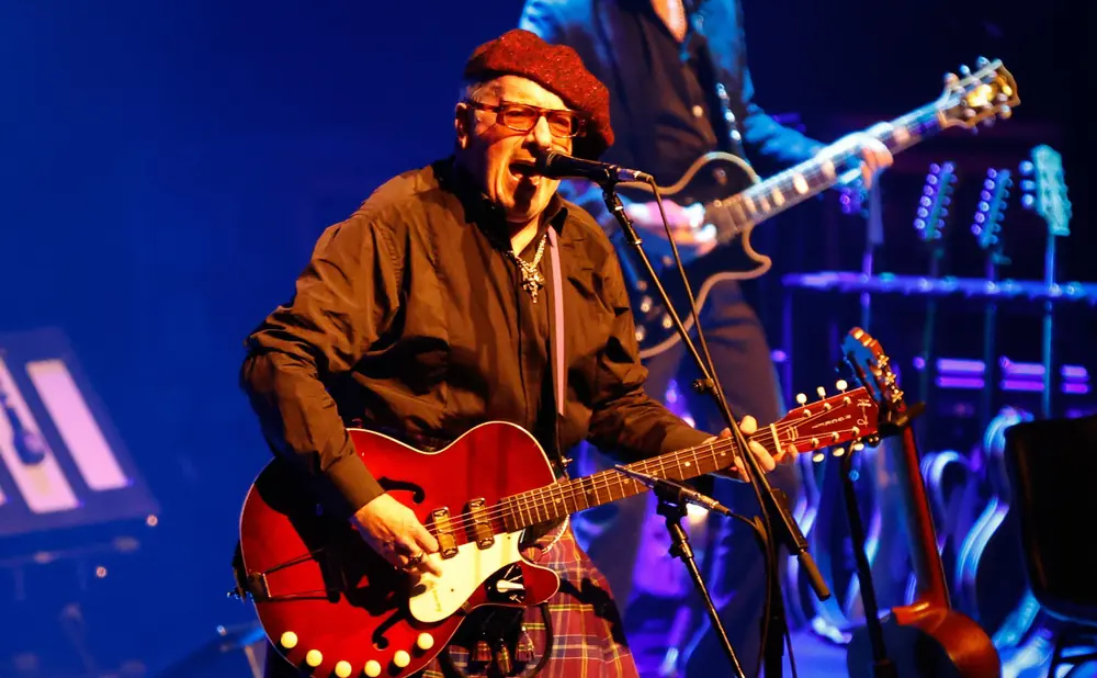 A musician in a red beret and tartan kilt plays a red guitar on stage with blue lighting.