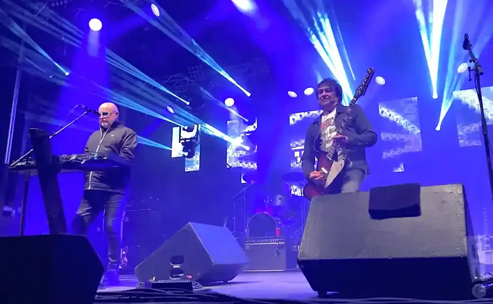 Two musicians performing on a brightly lit blue stage. One plays a keyboard, the other a guitar.