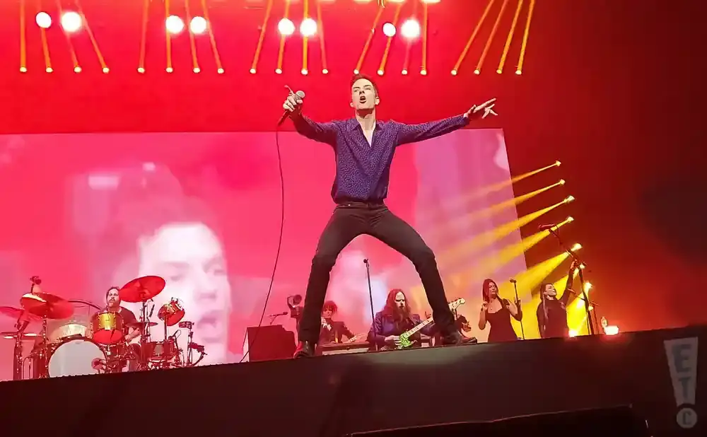 A male singer in a purple shirt and black pants performs on a stage with a large pink screen behind him.