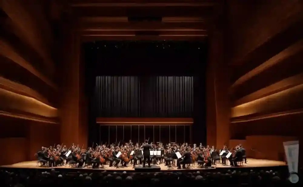 Orchestra performing on stage with a conductor, lit by warm stage lighting.