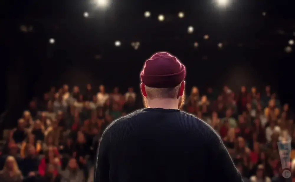 A person in a maroon beanie and dark sweater faces an audience from a stage, lit by spotlights.