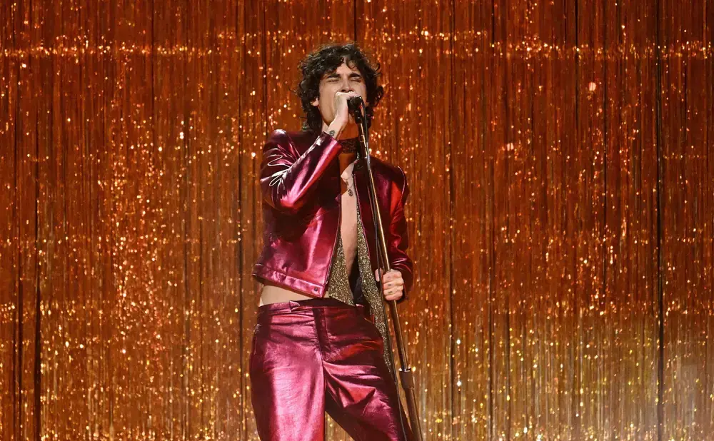 A singer in a sparkly red outfit performs on stage against a shimmering gold fringe curtain.