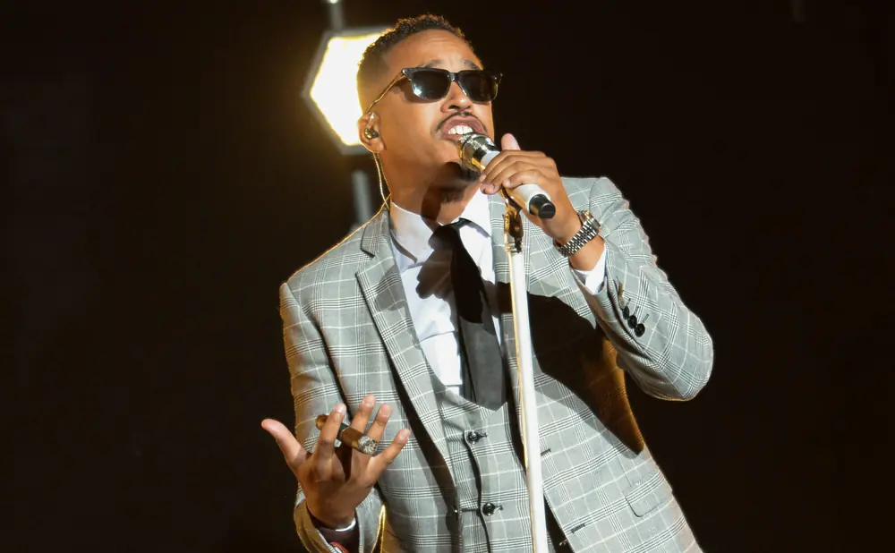 Man in a plaid suit and sunglasses sings into a microphone.