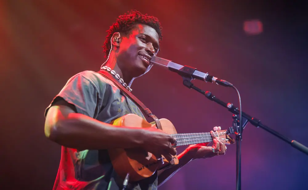 A Black man smiles while playing a ukulele and singing into a microphone on a stage with colorful lighting.