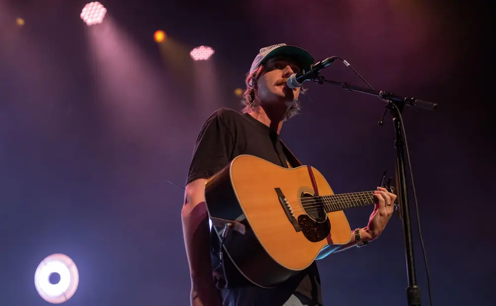 Musician in a cap sings into a microphone while playing an acoustic guitar.