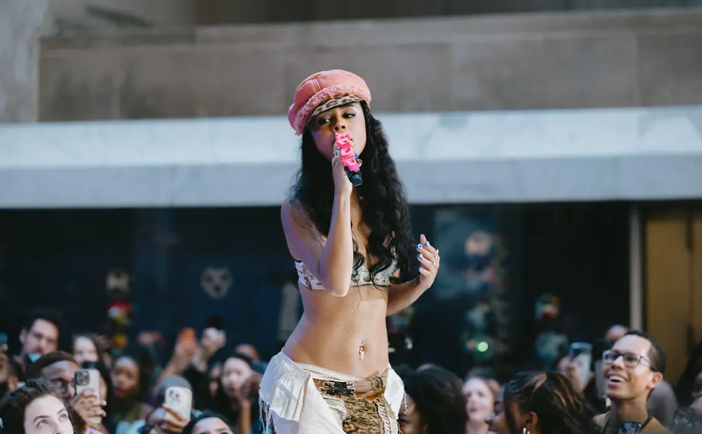 A female performer in a pink hat sings into a pink microphone in front of a blurred crowd.