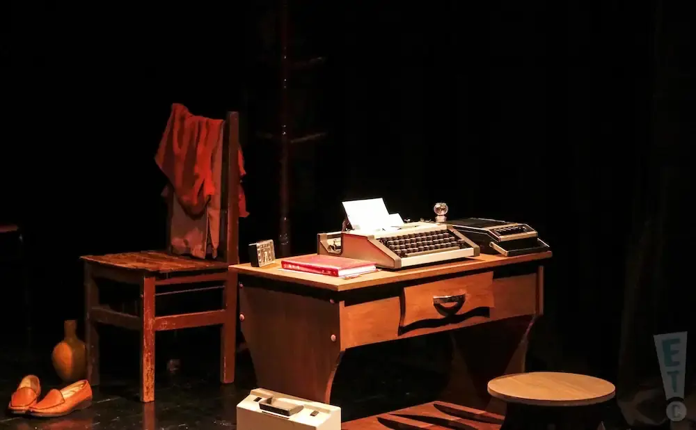 A staged desk with a typewriter and a chair with a red cloth draped over it.