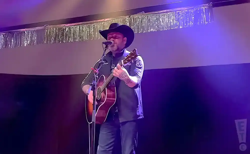 A man in a cowboy hat plays an acoustic guitar on stage, illuminated by purple lights.