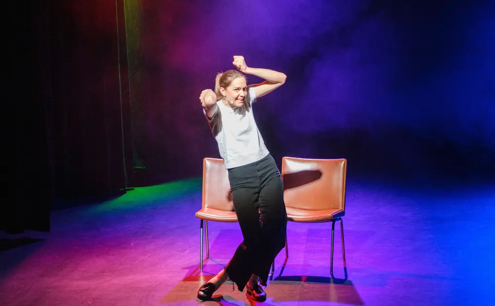 A dancer in black pants and a white shirt smiles as she strikes a pose on a stage with colorful lighting.