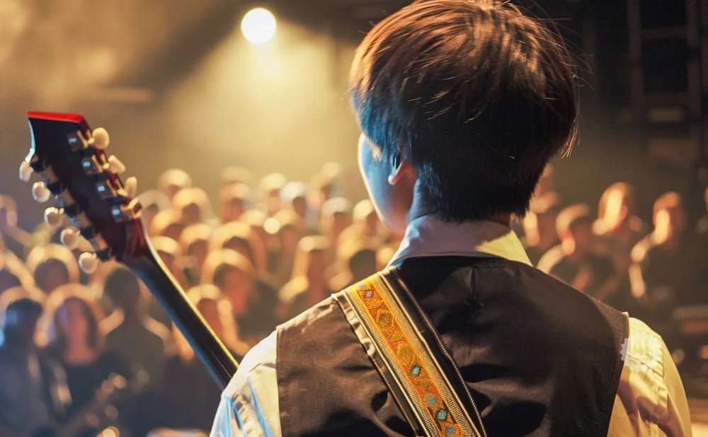 Musician playing guitar on stage with a blurred crowd in the background.