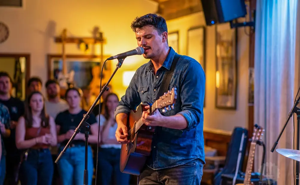 A male musician in a denim shirt sings and plays an acoustic guitar on stage in front of a blurred audience.