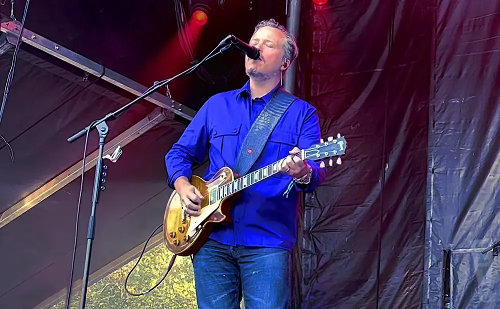 A man in a blue shirt plays a guitar and sings into a microphone on stage.