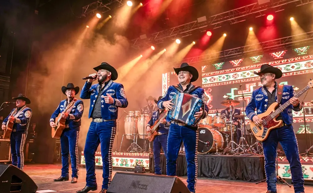 A band in matching blue sequined outfits and cowboy hats performs on stage with guitars, an accordion, and drums.