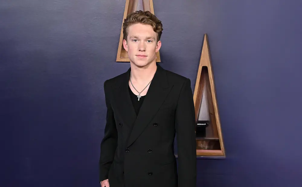 A young man in a black suit and a silver cross necklace poses against a purple wall with triangular wooden decor.