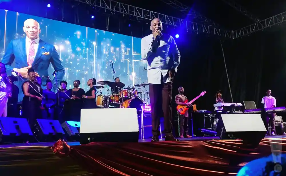 A man in a blue suit stands on stage singing as a band and choir perform behind him.