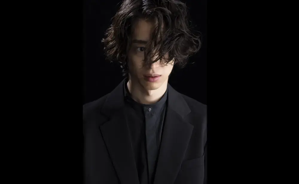 Man with dark, messy hair and a black suit jacket over a black shirt, looking forward against a black background.