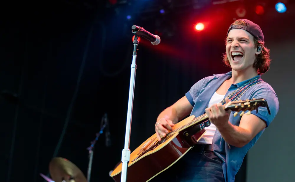 A male guitarist in a blue shirt and jeans sings passionately into a microphone on stage.