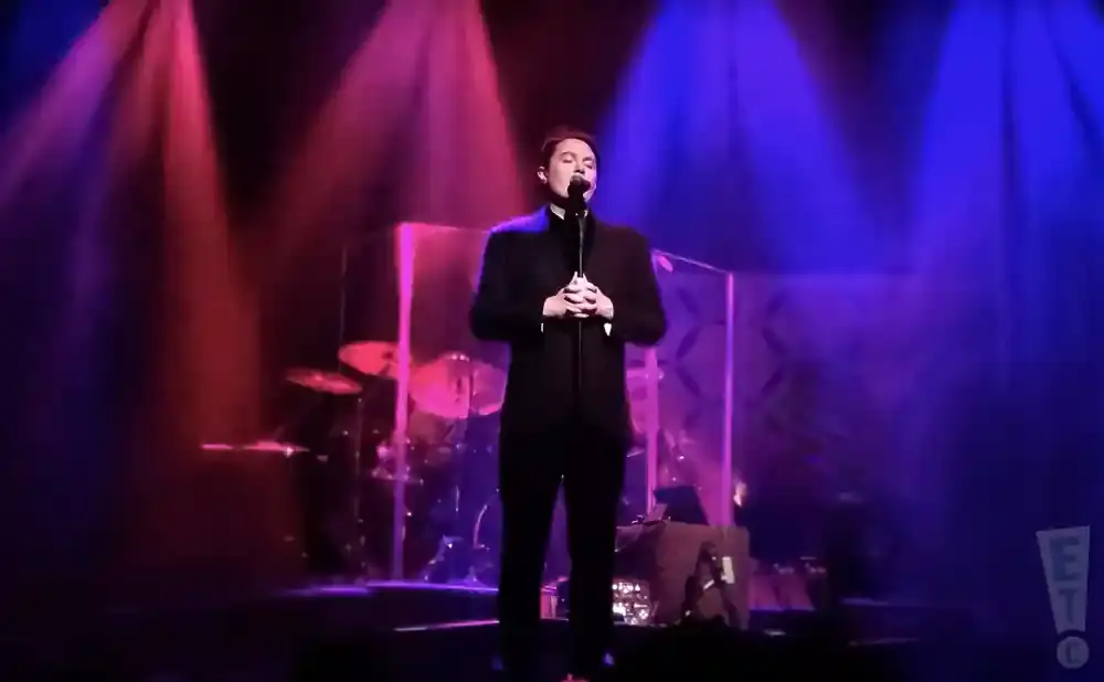 Man in a black suit sings into a microphone on a stage with red and blue lights.