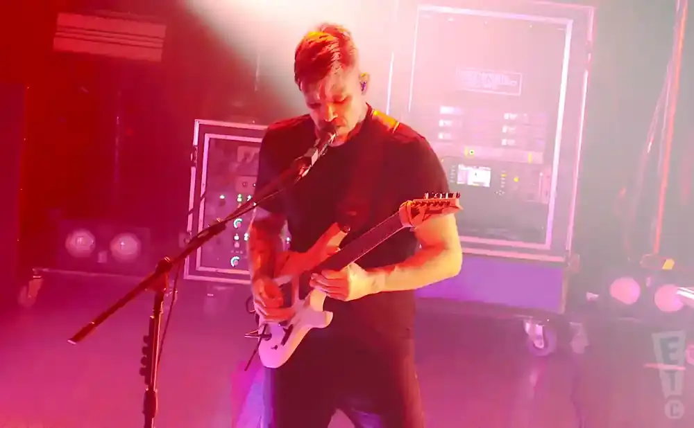 A guitarist plays a white electric guitar on a stage lit with pink and red lights.