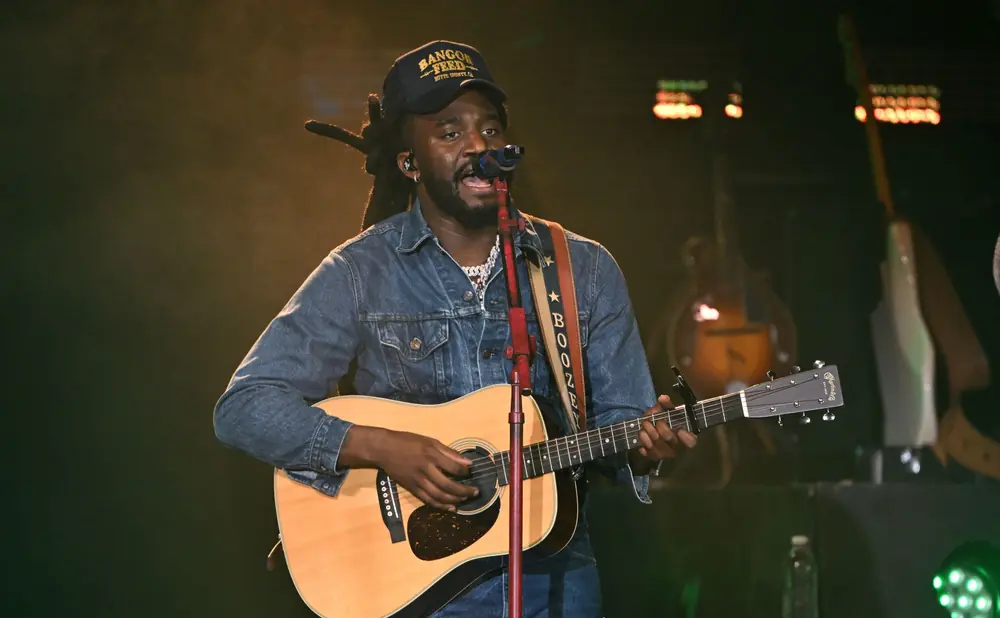 A Black man with dreadlocks wears a denim jacket and hat while playing an acoustic guitar on stage.