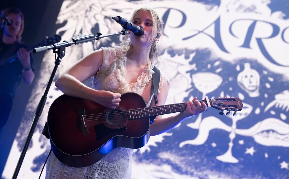 Woman with blonde hair plays guitar and sings into a microphone on stage with a blue and white patterned backdrop.