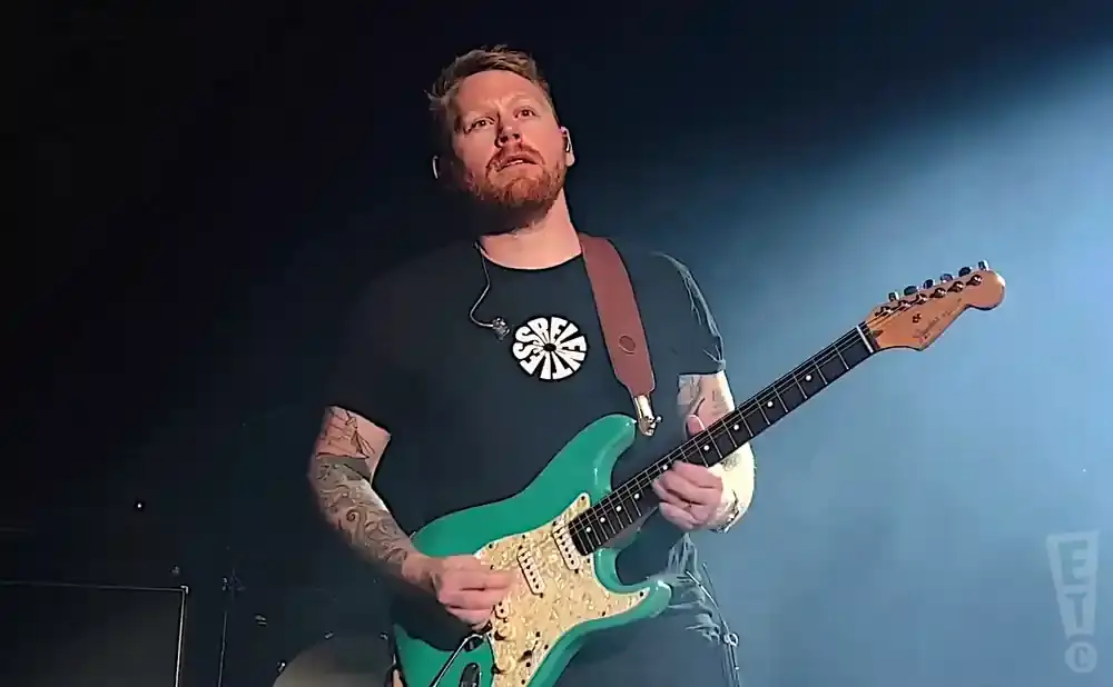 Guitarist with red beard and tattoos plays a teal electric guitar on stage.
