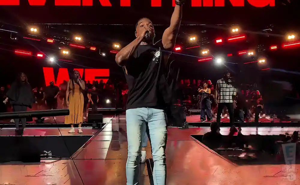 A male singer with his arm raised, performing on stage with a choir behind him.