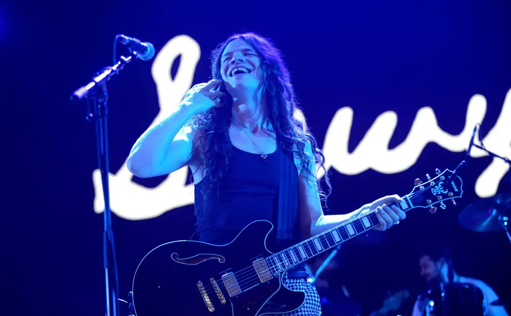 A male guitarist with long hair laughs on stage, holding a black Gibson guitar.