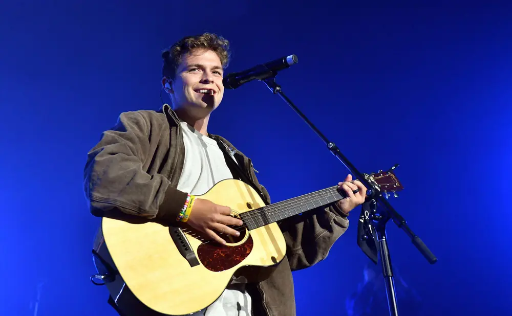 A man plays an acoustic guitar and sings into a microphone on a blue stage.
