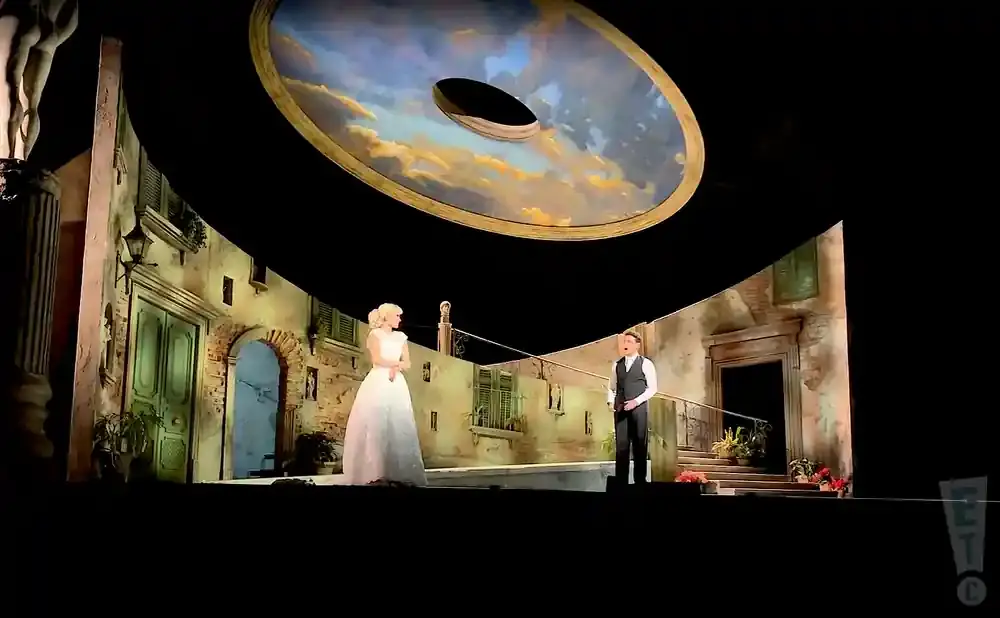 A stage with a painted sky backdrop, two singers in opera attire stand on a set of an old Italian street.