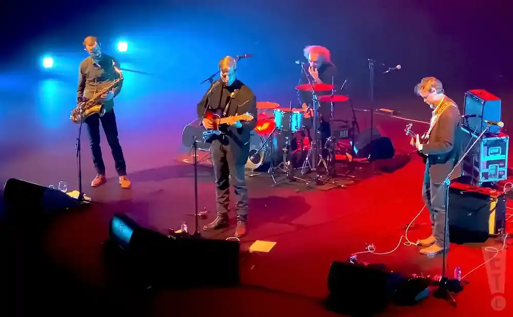Musicians perform on a stage with blue and red lights. A saxophonist, guitarist, and drummer are visible.