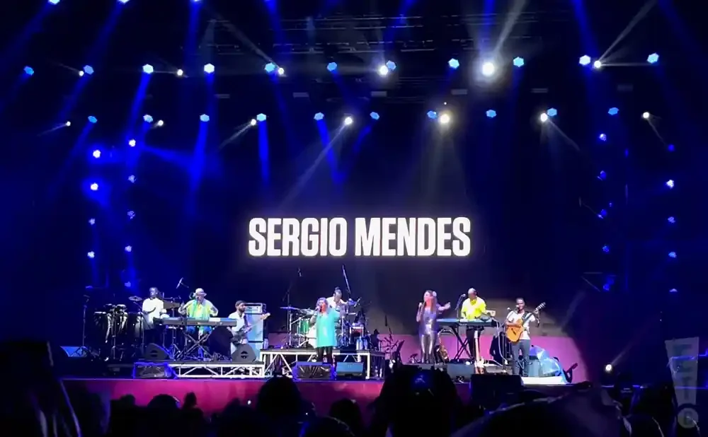 Sergio Mendes performing live on stage with his band, bathed in blue stage lights.