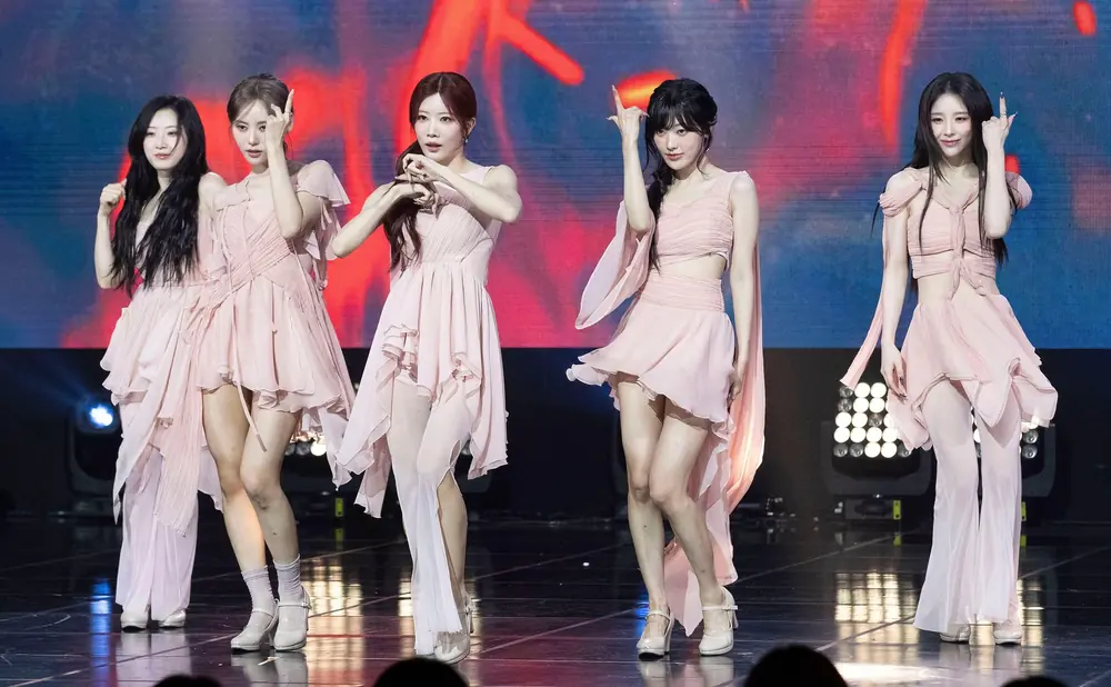 Four young women perform on stage, two playing bass guitars, one singing, and one with colorful hair smiling.