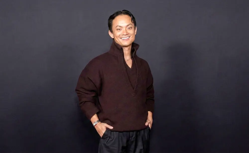 A man smiles at the camera, hands in his pockets, wearing a dark brown sweater and black pants.