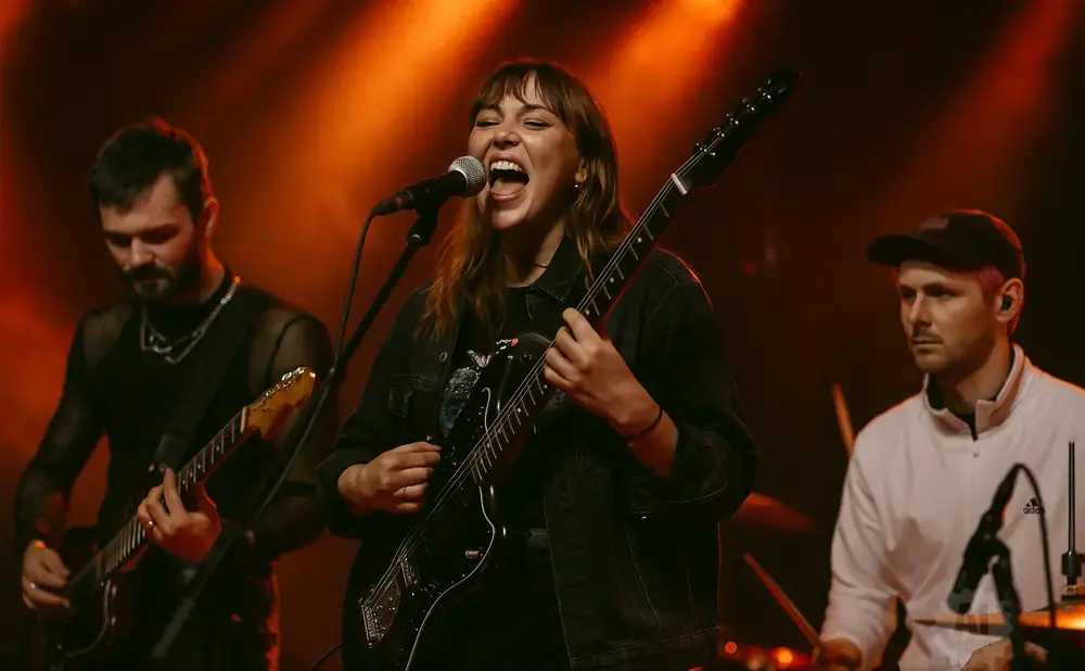 A band performs on stage with warm lighting. A woman sings and plays guitar center, flanked by a guitarist left and drummer right.