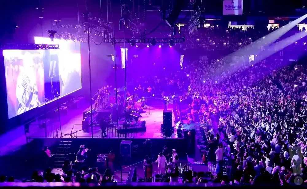 A band performs on a brightly lit stage with a large screen behind them, to an enthusiastic crowd in a stadium.