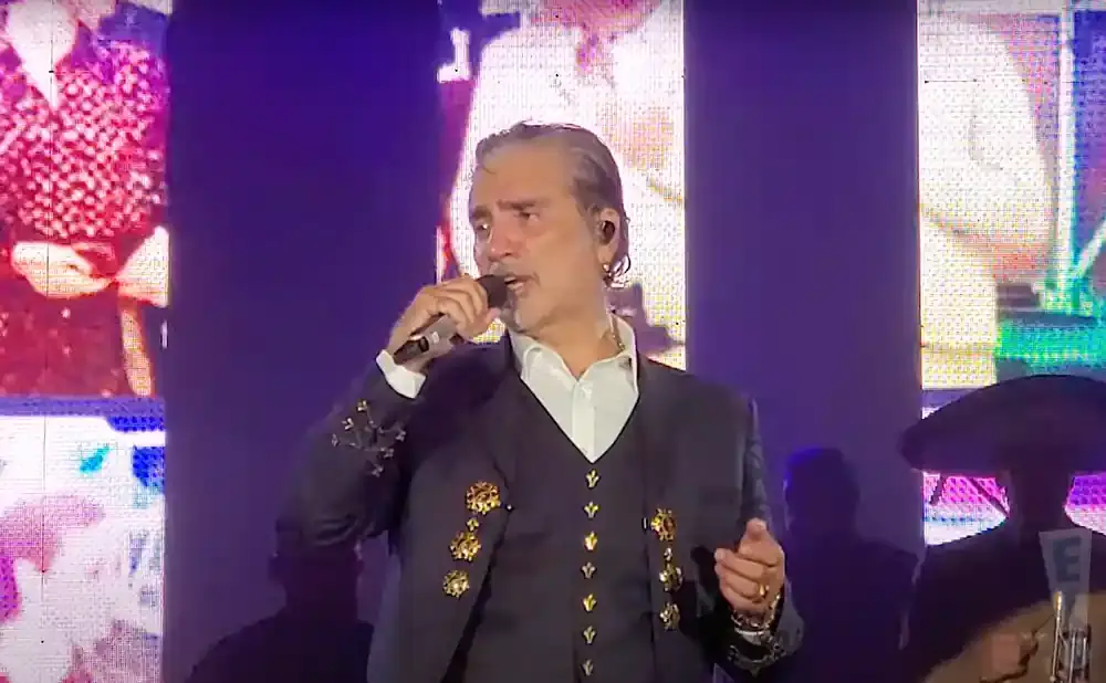 A man in a suit sings into a microphone on a stage with a colorful screen behind him.