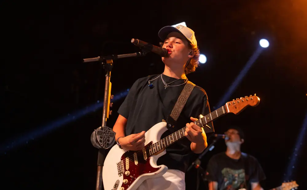 A young guitarist sings into a microphone while playing a white electric guitar on a dark stage.