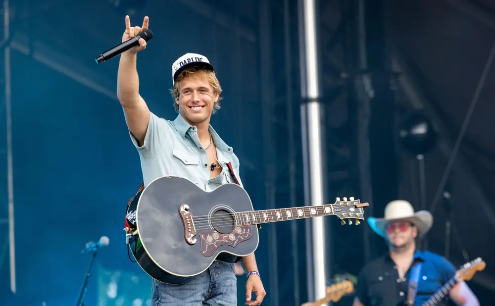 A male country singer with a guitar on stage holds a microphone and wears a baseball cap.