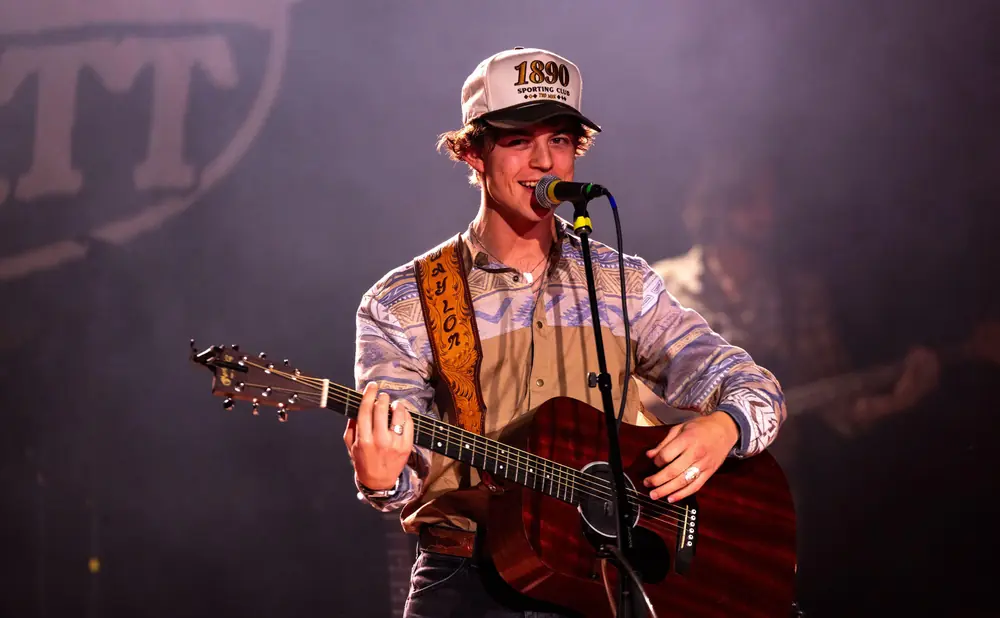 A young man with curly brown hair and a baseball cap sings and plays an acoustic guitar on stage.
