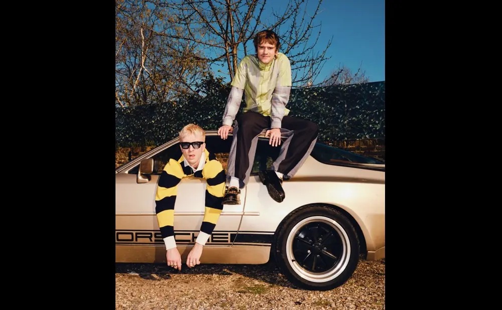 Two men pose by a Porsche. One leans out the window, the other sits on the roof.