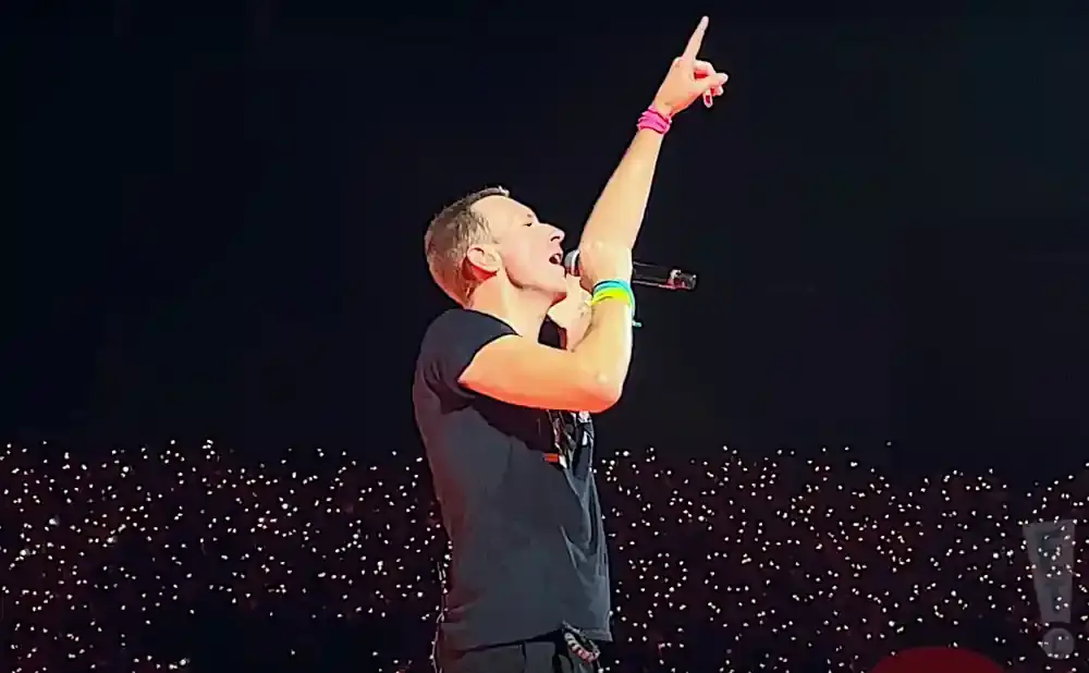 A man sings into a microphone with his arm raised and finger pointing up, on a stage in front of a crowd holding lights.