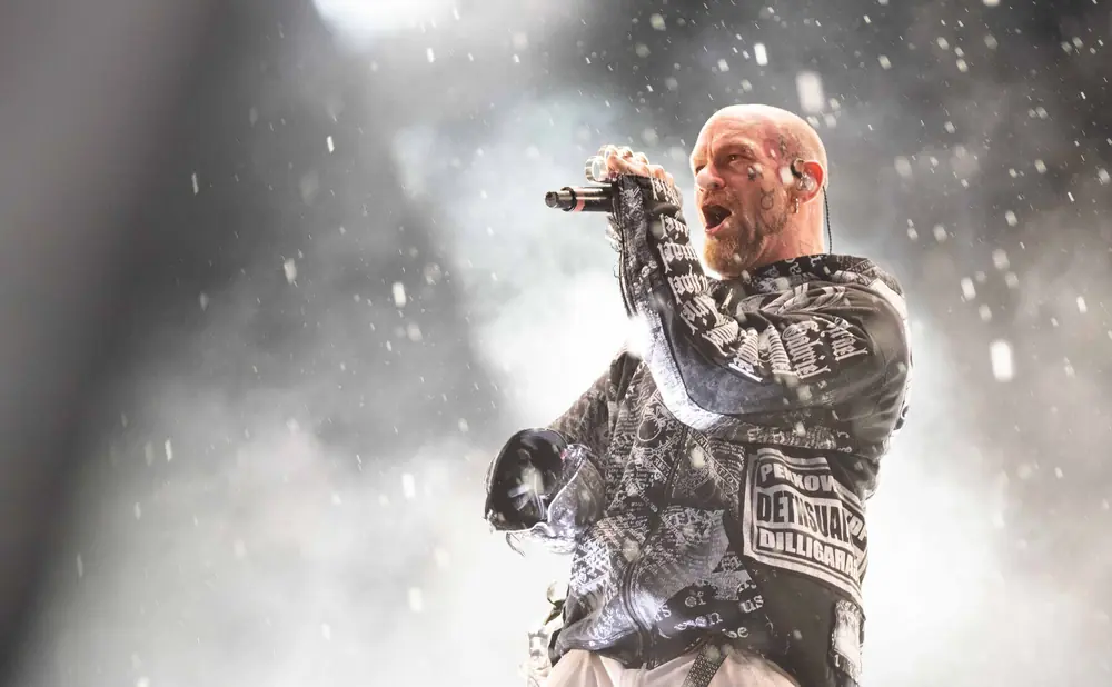 A bald man with tattoos on his face sings into a microphone on a smoky stage.