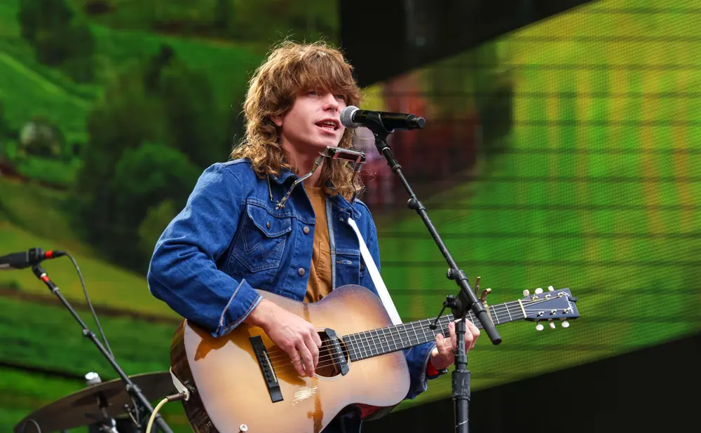 Man with long hair wearing a denim jacket plays an acoustic guitar and sings into a microphone at an outdoor concert.